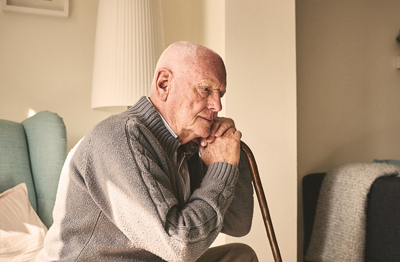 An elderly gentleman sat down using his walking stick to rest his head on his hands whilst looking into the distance