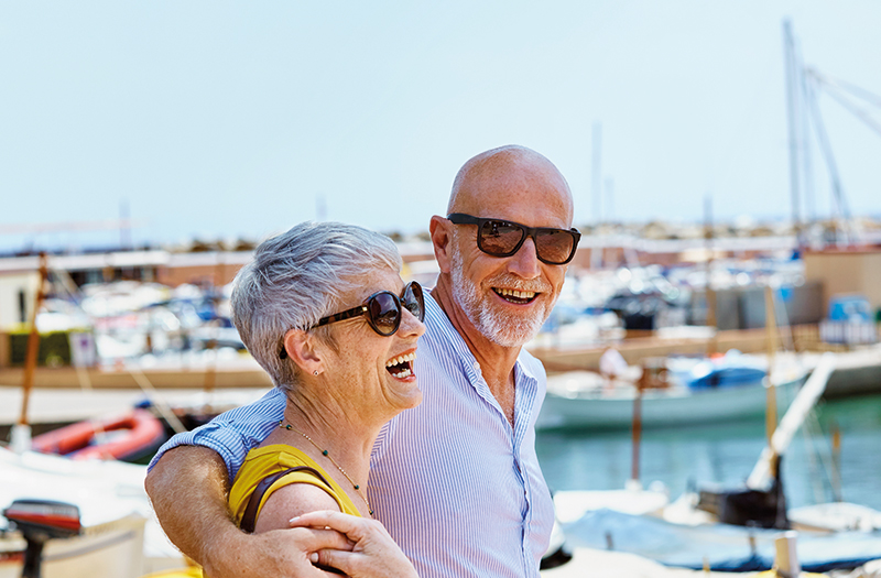 Elderly couple walking with their arms around each other smiling in their sunglasses