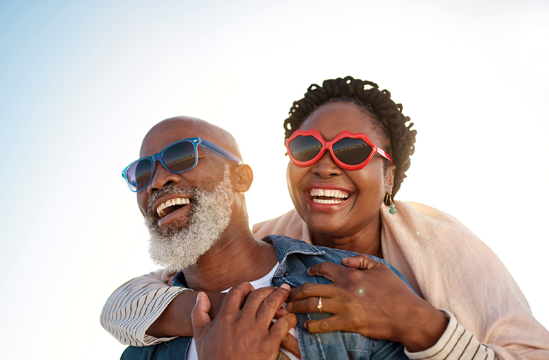 Elderly gentleman giving a piggyback to an elderly female as they both laugh in their sunglasses