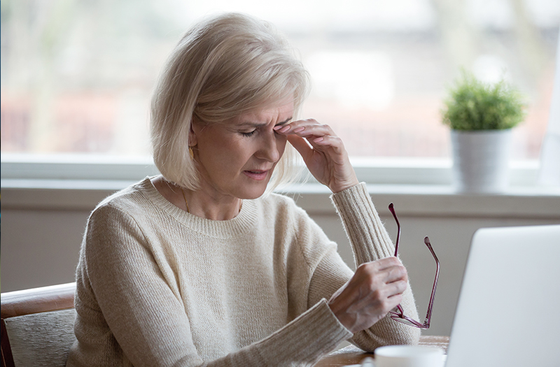 Elderly woman rubbing her sore eyes with one hand whilst holding her glasses in the other hand