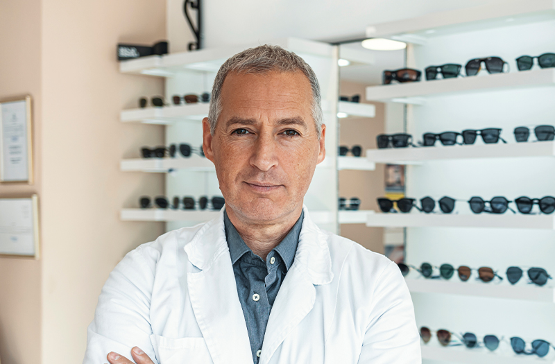 Happy optician stood with his arms crossed surrounded by a wide range of different glasses frames