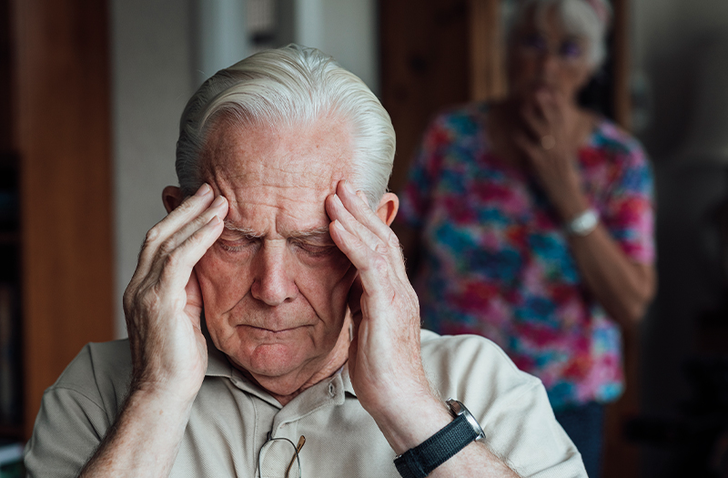 An elderly gentleman holding his head in his hands whilst a woman stands behind him with her hand to her mouth looking worried