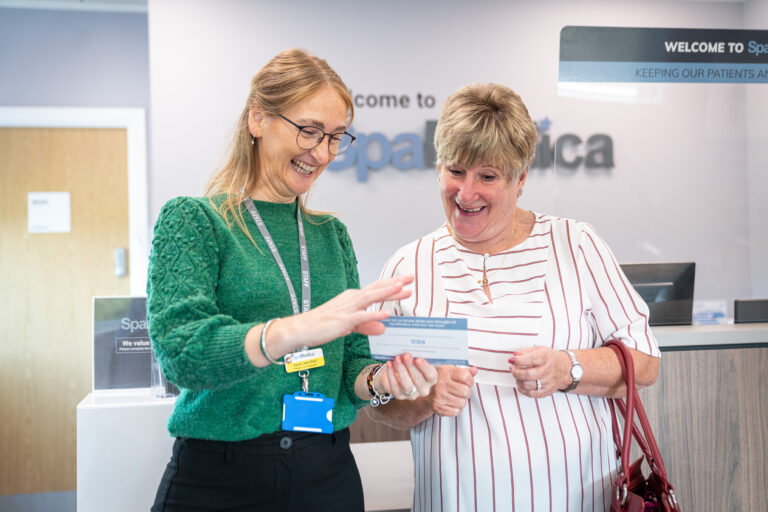 A smiling SpaMedica Patient Co-ordinator handing a feedback leaflet to a happy SpaMedica female patient