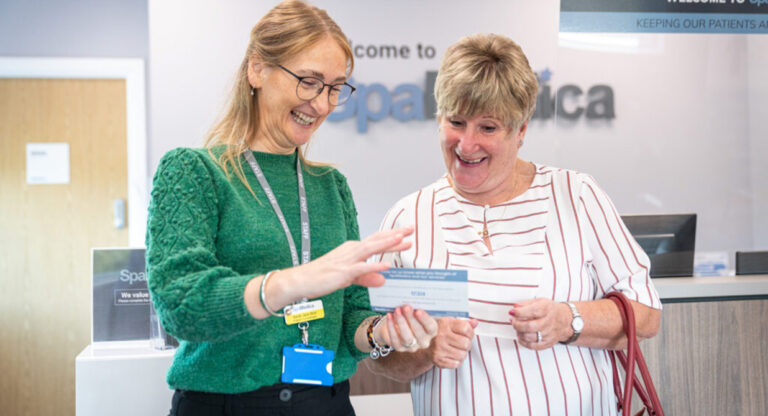 A smiling SpaMedica Patient Co-ordinator handing a feedback leaflet to a happy SpaMedica female patient