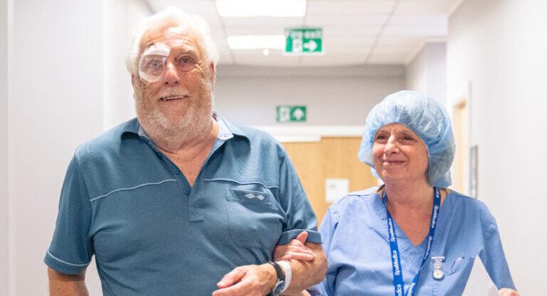 A happy SpaMedica patient, wearing an eye shield, linking arms and walking with a SpaMedica employee after successfully undergoing cataract surgery