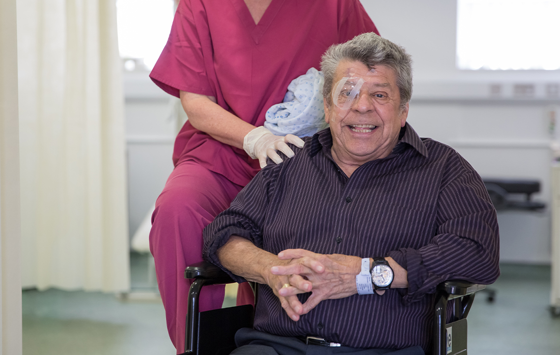 Delighted SpaMedica patient with eye shield post surgery being supported in a wheelchair by a SpaMedica employee