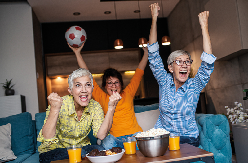 3 ecstatic women watching football on the couch and drinking orange juice